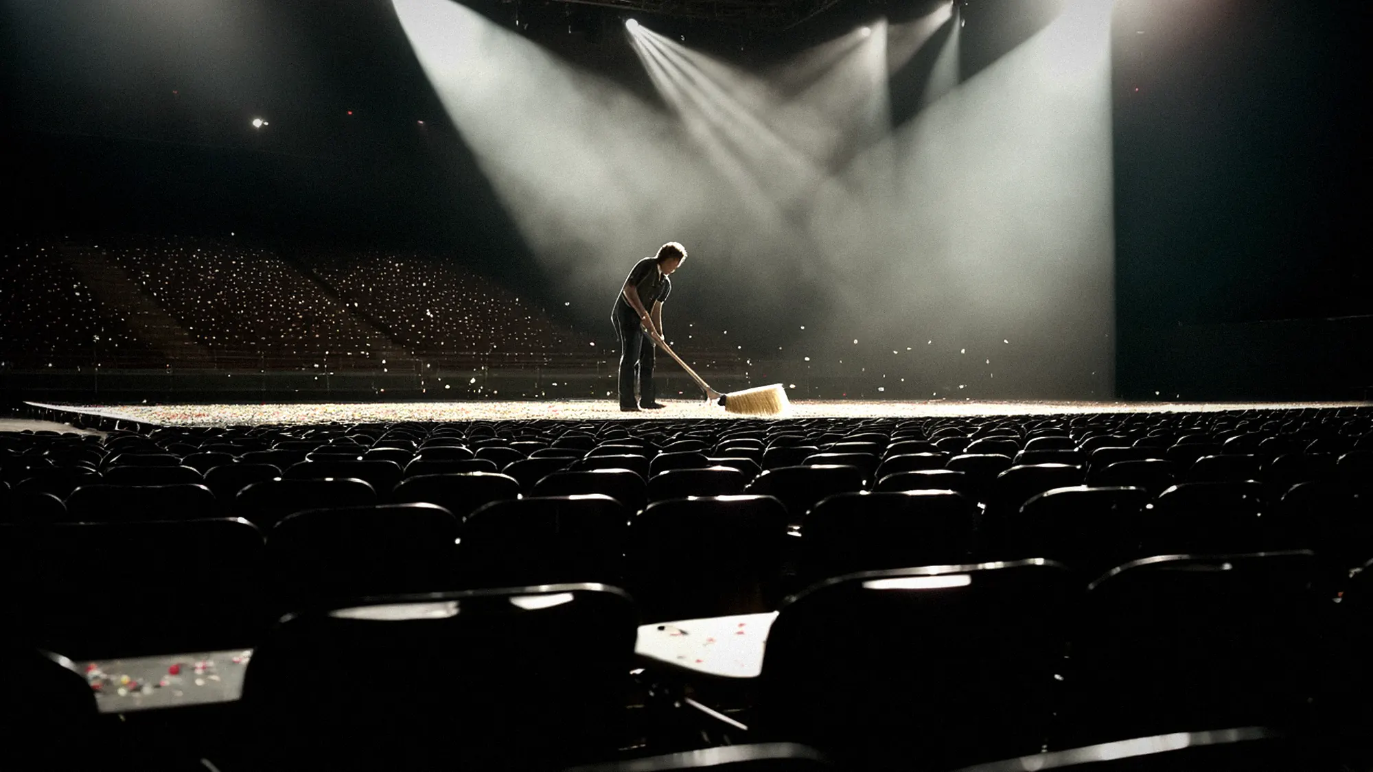 Leere Arena nach dem Konzert im Arbeitslicht, Sitzreihen und Konfetti, eine Person fegt in der Ferne